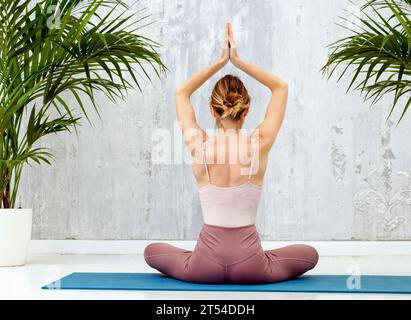 Rear view of a woman practicing the Sukhasana yoga pose sitting cross-legged on the floor with hands raised for relaxation, meditation and body alignm Stock Photo
