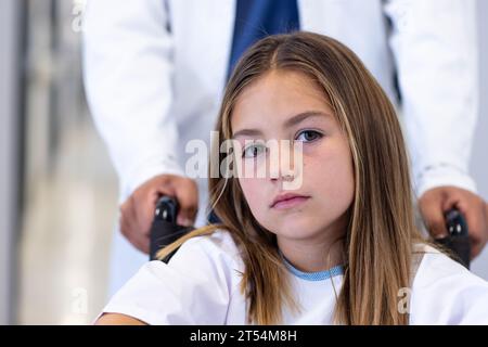 Portrait of caucasian girl patient in wheelchair being pushed by male doctor in hospital corridor Stock Photo