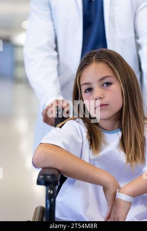 Portrait of caucasian girl patient in wheelchair being pushed by male doctor in hospital, copy space Stock Photo