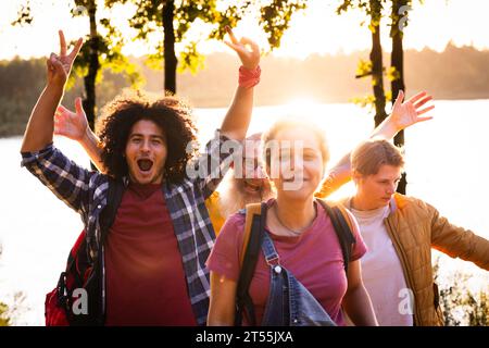 Bathed in the soft light of sunset, a scene of genuine camaraderie unfolds. Four multiracial millennials, their backpacks hinting at shared journeys, come together to celebrate in nature's embrace. The leading lady's smile, bright and infectious, captures the group's shared joy, while her friends in the background elevate the scene with their exuberant cheers. It's a snapshot of youth, friendship, and the simple pleasures of life. Golden Moments: Cheering Multiracial Millennial Friends in Nature. High quality photo Stock Photo