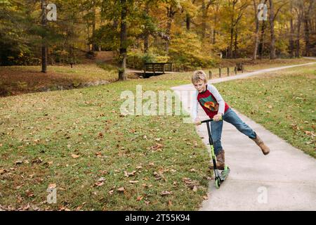 Boy riding scooter on sidewalk at a park during fall Stock Photo