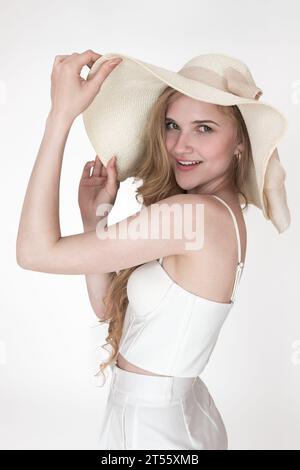 Portrait of fashionable Caucasian woman with raised hands holding brim of straw hat. Headshot smiling young adult female well dressed Stock Photo