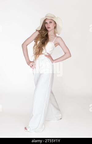 Full length photo of young blonde woman with wavy long hair wearing sun hat, white sculpting cupped corset top and trousers standing white background Stock Photo