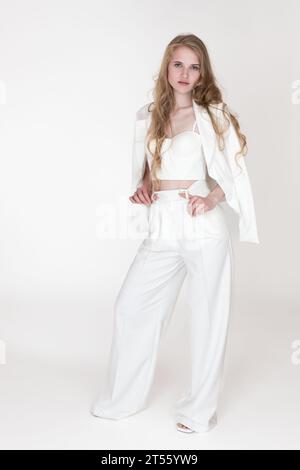 Well dressed Caucasian woman with blonde long wavy hair wearing white suit jacket draped over shoulders, sculpting cupped corset top, pants and sandal Stock Photo