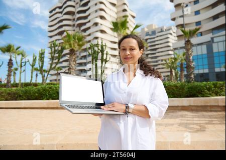 Attractive woman real estate agent holding laptop and showing copy space on white blank screen mockup, standing against modern high rise buildings bac Stock Photo