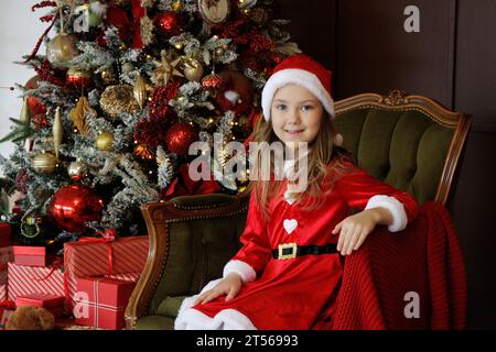 Little girl in santa dress sitting on green armchair near christmas tree Stock Photo