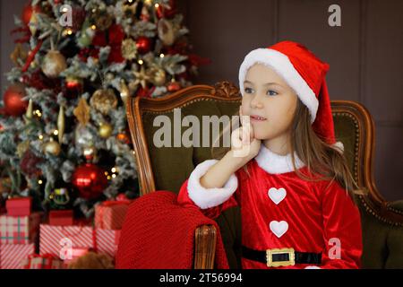 Thinking little girl in santa dress sitting on green armchair near christmas tree Stock Photo