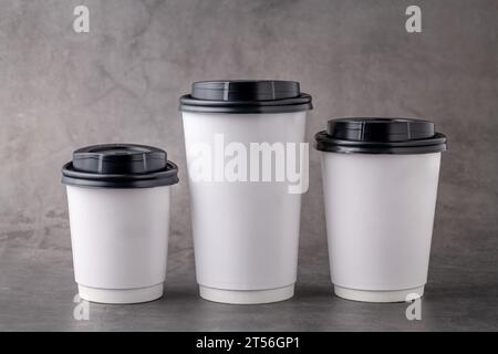 Three different sizes of white takeaway paper cups with black plastic lids isolated on gray background. Stock Photo