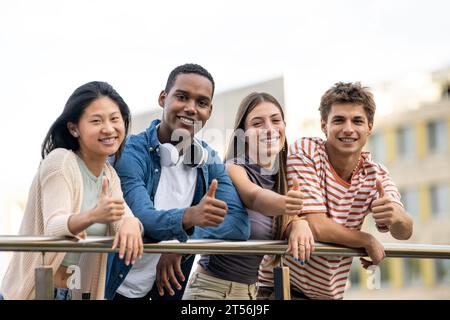 Beautiful group of friends satisfied and looking at camera leaning on a balcony. Diverse group of cheerful and hopeful millennial people showing thumb Stock Photo