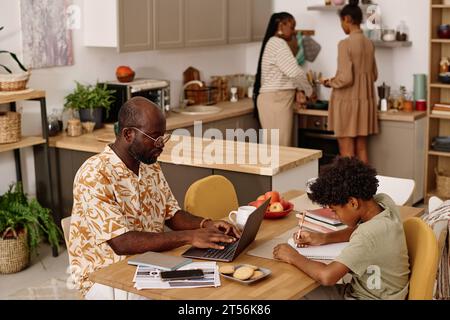 Father working on laptop, son doing homework when mother and daughter cooking dinner Stock Photo