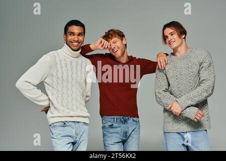 cheerful diverse male models in casual sweaters laughing and posing on gray backdrop, men power Stock Photo