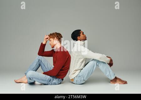 two young multiethnic men in cozy warm sweaters sitting on floor back to back, fashion concept Stock Photo