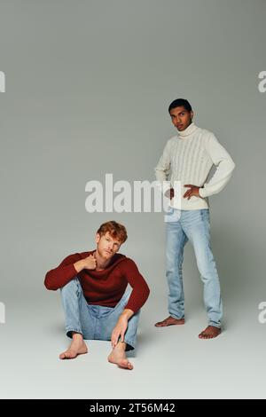 red haired man in sweater squatting on floor with african american man standing on gray background Stock Photo