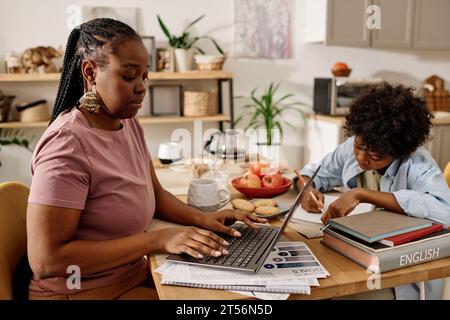 Black woman working on laptop at home when her son doing homework Stock Photo