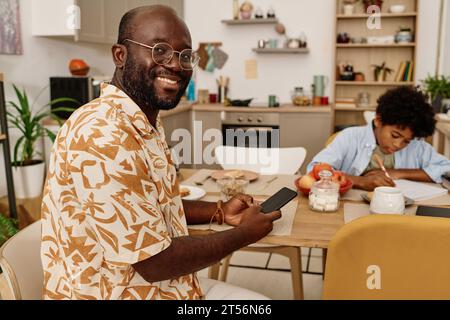 Smiling Black man answering text messages from colleagues when his son doing homework Stock Photo