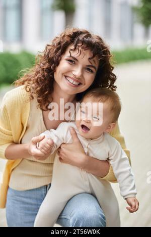 happy woman with wavy hair smiling at camera near little daughter outdoors, family portrait Stock Photo