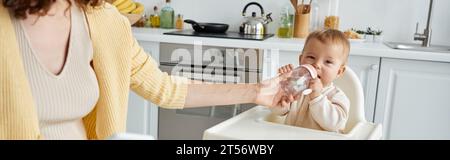 caring mother helping toddler girl drinking from baby bottle in kitchen, banner Stock Photo
