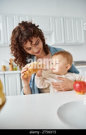 curly woman giving delicious croissant to toddler daughter near fresh apple, breakfast in kitchen Stock Photo