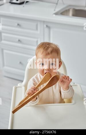 funny toddler girl sitting in baby chair and chewing wooden cooking tongs, morning in kitchen Stock Photo