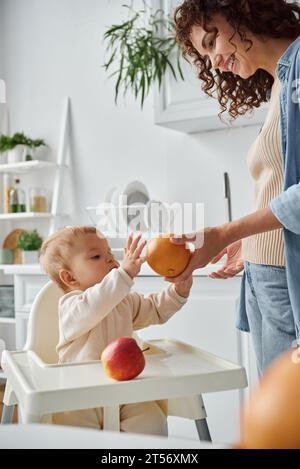 smiling mother giving fresh orange to little daughter in baby chair in kitchen, healthy breakfast Stock Photo
