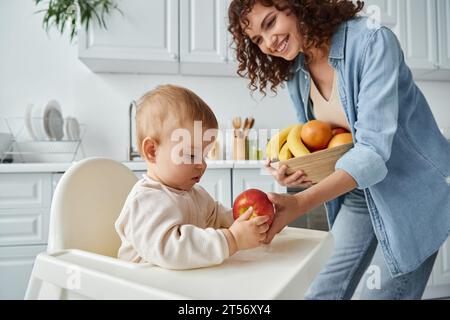 cheerful woman giving ripe apple to toddler daughter sitting in baby chair in kitchen, breakfast Stock Photo