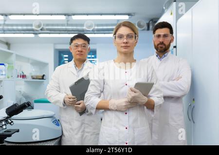 Team of doctors, meditators, lab assistants in white medical coats, serious diverse people looking at camera, holding tablet computer in hands working inside laboratory. Stock Photo