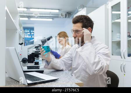 Serious and thoughtful scientist working in laboratory with microscope and laptop, mature experienced man in glasses and medical gown conducting experiments and entering data online into computer. Stock Photo