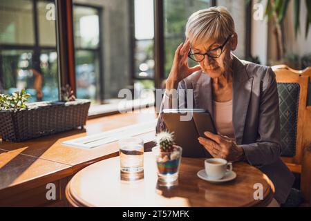 Mature woman is sitting in cafe and relaxing. She is drinking coffee and using digital tablet. Stock Photo