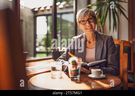 Mature woman is sitting in cafe and relaxing. She is drinking coffee and using note pad. Stock Photo
