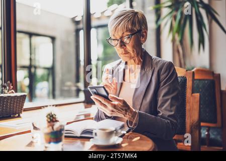 Mature woman is sitting in cafe and relaxing. She is drinking coffee and messaging on phone. Stock Photo