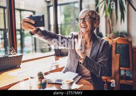 Mature woman is sitting in cafe and relaxing. She is drinking coffee and using smartphone. Stock Photo