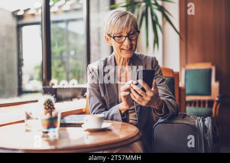 Mature woman is sitting in cafe and relaxing. She is drinking coffee and messaging on phone. Stock Photo