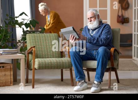 Senior man sitting on armchairs and using tablet pc at home with his wife in background Stock Photo