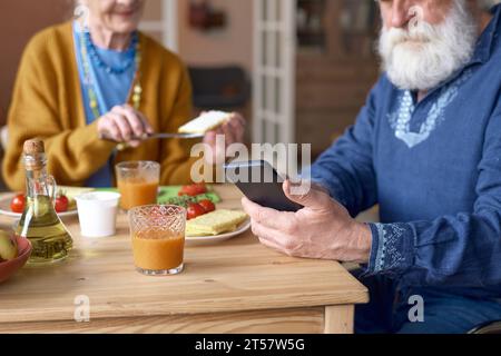 Close-up of senior man using smartphone while sitting at table and having lunch with his wife Stock Photo