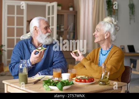 Senior couple eating sandwiches and talking to each other while having breakfast at home Stock Photo