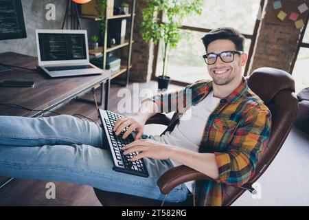 Profile photo of smart positive web designer guy sitting chair legs desk keyboard typing database optimization modern loft office indoors Stock Photo