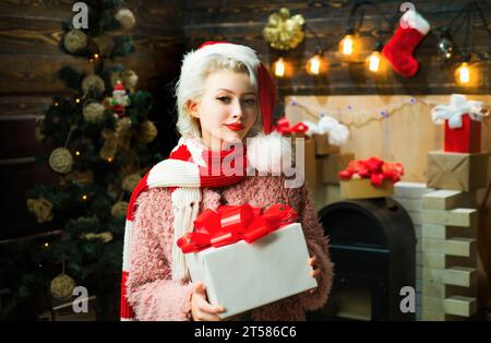 Blonde female model dressed in a Santa Claus hat. Cute young woman with santa hat. Euphoria. Fashion portrait of girl indoors with Christmas tree. Stock Photo