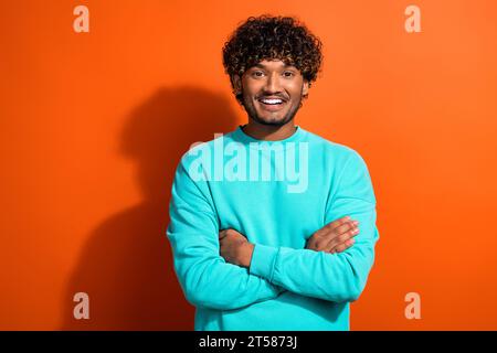 Portrait of smiling guy wearing stylish blue sweatshirt folded arms enjoying his new curly hairstyle isolated on orange color background Stock Photo