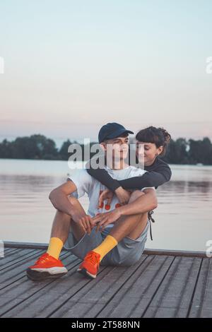 Cute young couple on the shore of a lake looking into their future during sunset. 20-year-old couple hugging. Stock Photo