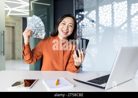Portrait of young joyful asian business woman, woman smiling and looking at camera, holding phone and winning money cash american dollars, working at workplace inside office. Stock Photo
