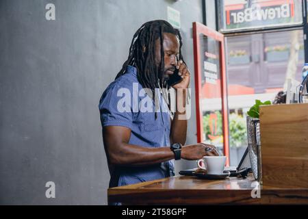 Side view of black businessman, with dreadlocks, beard and casual clothing, on phone call using laptop standing inside restaurant, concentrated listen Stock Photo