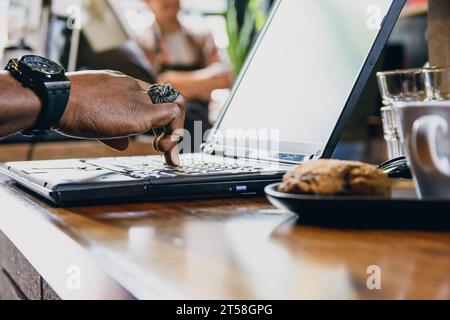 side view hand of unrecognizable black man touching with one finger laptop keyboard on table inside restaurant, with cookie and coffee cup out of focu Stock Photo