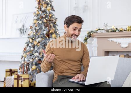 A young man is talking on a video call on a laptop while sitting on the sofa at home. Smiling and gesturing to the camera. Stock Photo