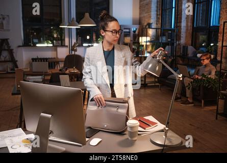 Serious young mixed race businesswoman in glasses standing with bag at table and turning off light in office Stock Photo