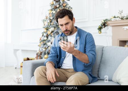 Upset unhappy man sitting near Christmas tree on winter day, got bad news on phone, celebrating new year and Christmas, unhappy reading, using app on smartphone, living room at home on sofa. Stock Photo