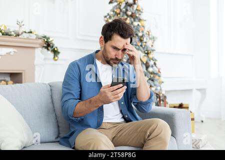 Upset unhappy man sitting near Christmas tree on winter day, got bad news on phone, celebrating new year and Christmas, unhappy reading, using app on smartphone, living room at home on sofa. Stock Photo