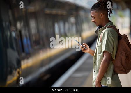 Young Man Commuting To Work On Train Standing  On Platform Looking At Mobile Phone As Train Arrives Stock Photo
