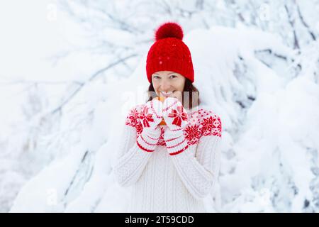 Woman drinking hot chocolate in Christmas morning in snowy garden. Girl in knitted Nordic sweater, hat and mittens holding cup with cocoa. Stock Photo