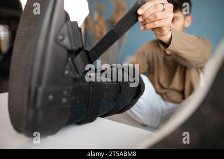 Man putting ankle sprain orthosis, black splint on leg sitting on the sofa. Ankle foot orthosis. Stock Photo