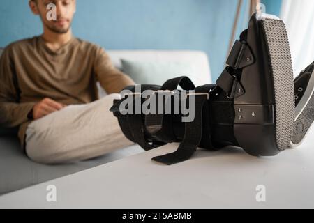 Young man with ankle orthosis, walker boot on leg sitting on the sofa at home, close-up. Stock Photo